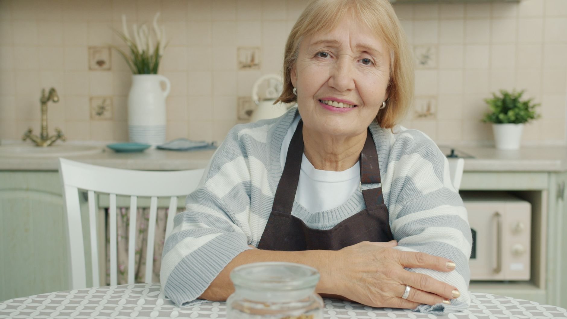 Elderly woman in apron sitting in a warm kitchen, smiling.